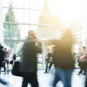 A motion-blurred image of a man speaking on a mobile phone while walking through a crowded but bright and airy indoor space.