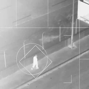 An infrared image of a person walking down a city street, overlaid with graphic elements from motion-tracking software including a white framing grid and a white square and circle superimposed over the person.
